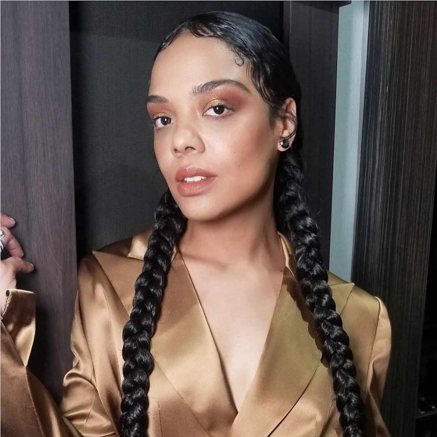 Tessa Thompson wears braided pigtails, a warm-toned makeup look, and a gold satin blazer