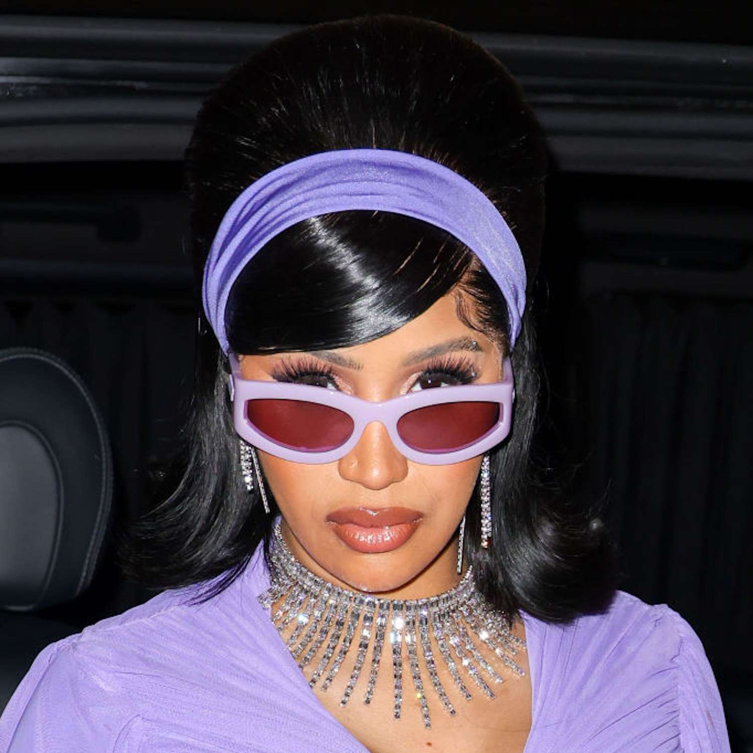 Cardi B with a bouffant and a purple headband, looking over her pink sunglasses